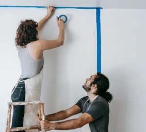 A couple engaged in a home renovation project, using a ladder and tape to prepare walls.