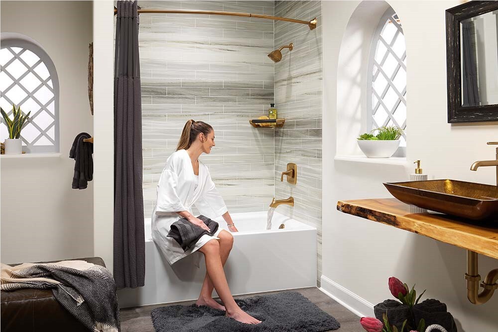 Photo of a woman sitting on the ledge of a bathtub and shower combo with acrylic wall panels and gold hardware